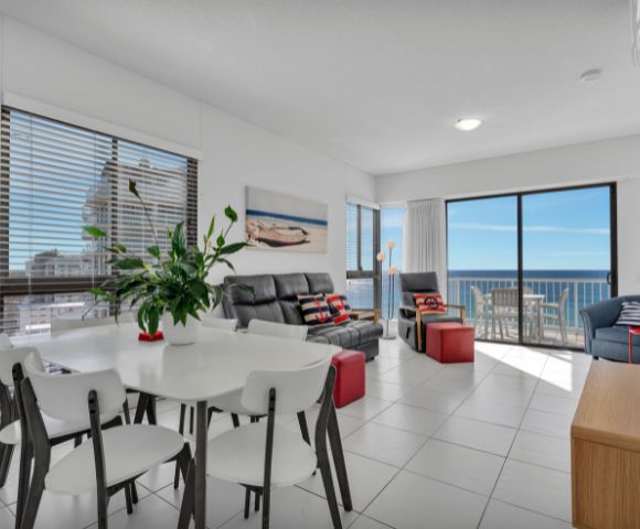 Bright, modern living room with ocean view. Features white walls, black sofas, red and blue accents, greenery, and glass doors leading to a sunny balcony.