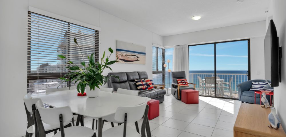 Bright, modern living room with ocean view. Features white walls, black sofas, red and blue accents, greenery, and glass doors leading to a sunny balcony.