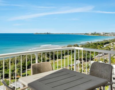 Balcony view of a bright coastal scene with a calm blue ocean, green shoreline, and a clear sky. Outdoor table and chairs suggest a relaxing atmosphere.