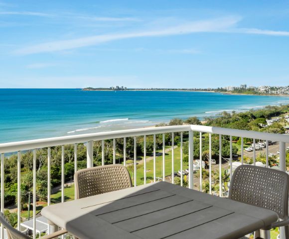 Balcony view of a bright coastal scene with a calm blue ocean, green shoreline, and a clear sky. Outdoor table and chairs suggest a relaxing atmosphere.