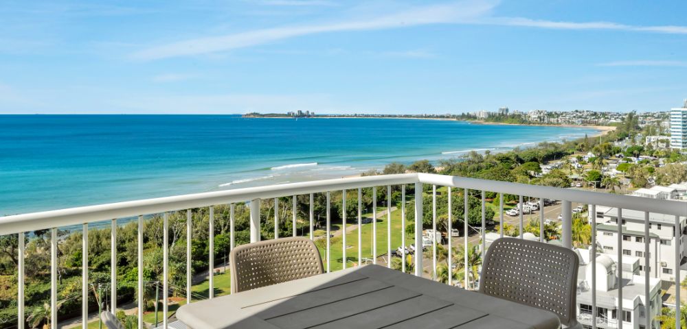 Balcony view of a bright coastal scene with a calm blue ocean, green shoreline, and a clear sky. Outdoor table and chairs suggest a relaxing atmosphere.