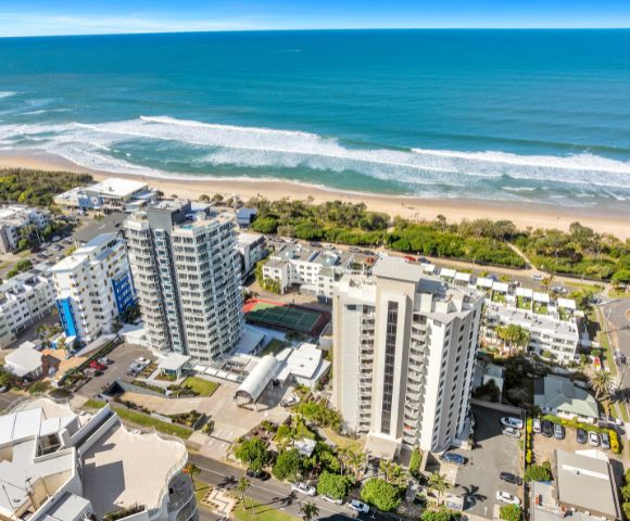 Aerial view of a coastal cityscape with tall buildings near a sandy beach and ocean waves under a clear blue sky, conveying a serene and vibrant atmosphere.