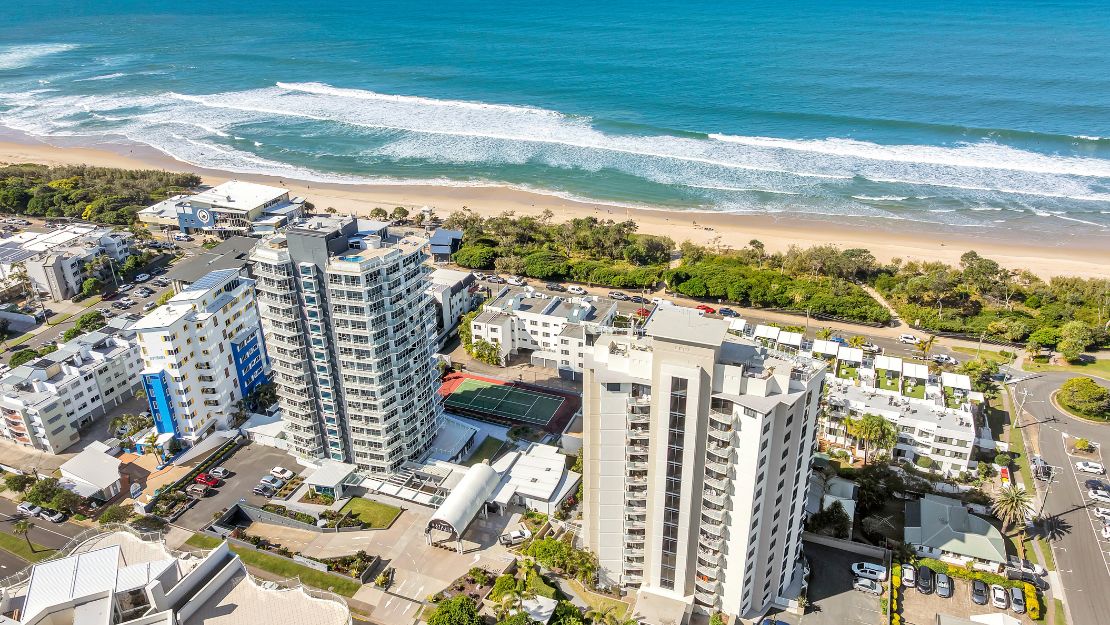 Aerial view of a coastal cityscape with tall buildings near a sandy beach and ocean waves under a clear blue sky, conveying a serene and vibrant atmosphere.