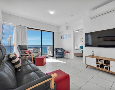 Modern living room with ocean view. Features black leather sofa, red ottoman, flat-screen TV, and glass doors opening to balcony. Bright, airy ambiance.
