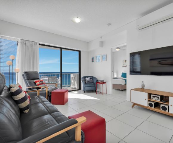 Modern living room with ocean view. Features black leather sofa, red ottoman, flat-screen TV, and glass doors opening to balcony. Bright, airy ambiance.