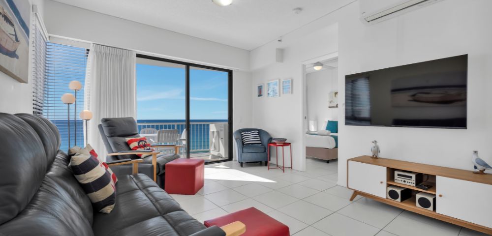 Modern living room with ocean view. Features black leather sofa, red ottoman, flat-screen TV, and glass doors opening to balcony. Bright, airy ambiance.