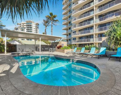 Outdoor pool with turquoise lounge chairs beside a high-rise building. Palm trees and clear blue sky add a tropical, relaxing atmosphere.