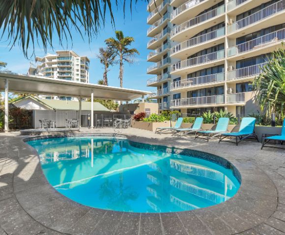 Outdoor pool with turquoise lounge chairs beside a high-rise building. Palm trees and clear blue sky add a tropical, relaxing atmosphere.
