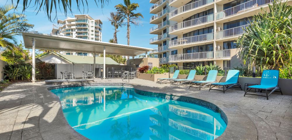 Outdoor pool with turquoise lounge chairs beside a high-rise building. Palm trees and clear blue sky add a tropical, relaxing atmosphere.