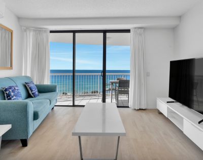 Modern living room with a teal sofa, cushions, and TV. Floor-to-ceiling windows showcase an ocean view and balcony with chairs, creating a serene atmosphere.