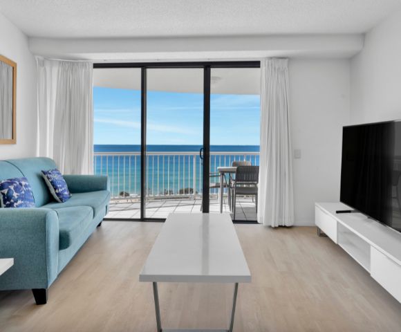 Modern living room with a teal sofa, cushions, and TV. Floor-to-ceiling windows showcase an ocean view and balcony with chairs, creating a serene atmosphere.
