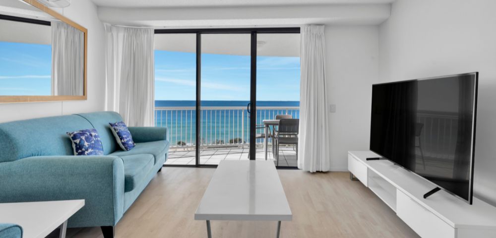 Modern living room with a teal sofa, cushions, and TV. Floor-to-ceiling windows showcase an ocean view and balcony with chairs, creating a serene atmosphere.