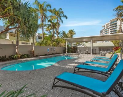 Outdoor pool area with turquoise lounge chairs on a tiled deck. Palm trees surround the pool, with city buildings visible in the background under a clear blue sky.