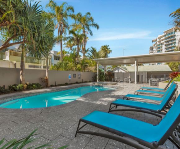 Outdoor pool area with turquoise lounge chairs on a tiled deck. Palm trees surround the pool, with city buildings visible in the background under a clear blue sky.