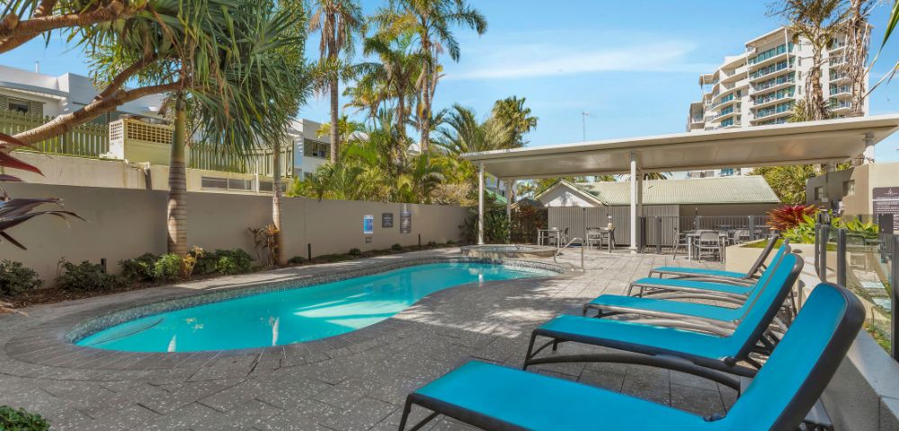 Outdoor pool area with turquoise lounge chairs on a tiled deck. Palm trees surround the pool, with city buildings visible in the background under a clear blue sky.