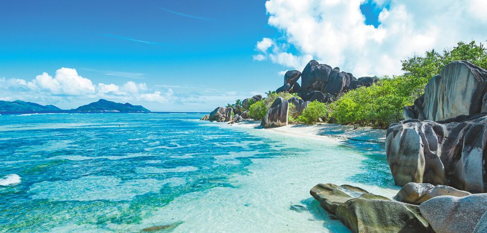 A serene tropical beach with turquoise water, smooth boulders, and lush greenery under a bright blue sky. A distant island is visible on the horizon.