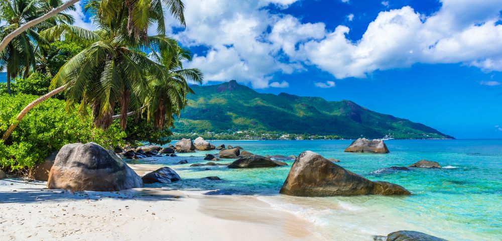 Tropical beach with turquoise waters, large rocks, and lush palm trees under a vibrant blue sky with fluffy clouds. Mountains loom in the background.