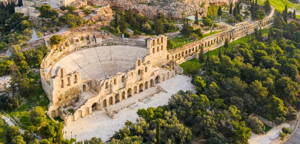 Aerial view of the ancient stone amphitheater, the Odeon of Herodes Atticus, nestled in lush greenery on a sunny day, conveying historical grandeur.
