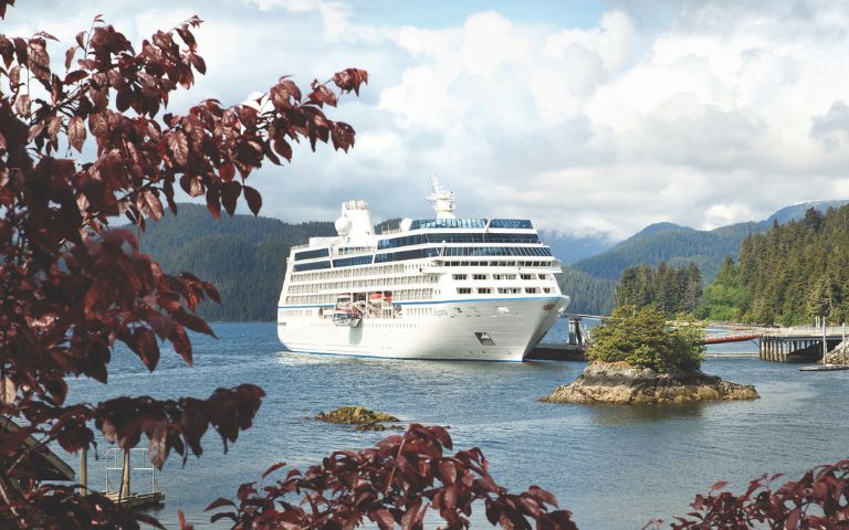 A large white cruise ship sails near a forested shoreline, with a small rocky island in the foreground and mountains under a cloudy sky in the background.