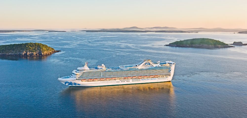 A large cruise ship glides through calm blue waters at sunset, surrounded by small, green islands. The scene conveys tranquility and adventure.
