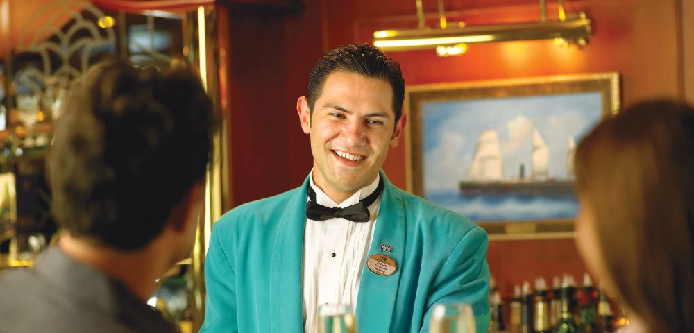 A smiling man in a teal jacket and black bow tie engages with two people in the foreground. A ship painting is blurred in the background, creating a warm and welcoming atmosphere.