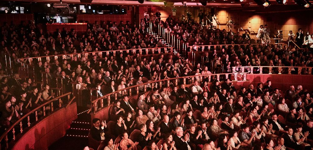 A large theater audience claps enthusiastically, filling the venue. Warm lighting and a sense of excitement surround the packed tiers of seated people.
