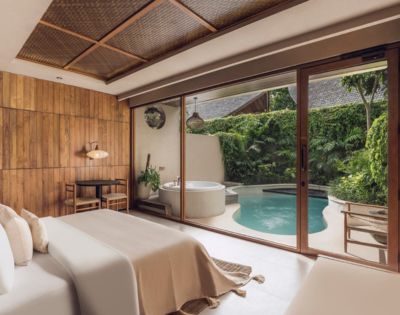 A serene bedroom with a wooden accent wall and soft beige bedding opens to a lush patio featuring a round jacuzzi and a small pool, surrounded by greenery.