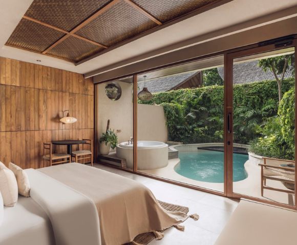 A serene bedroom with a wooden accent wall and soft beige bedding opens to a lush patio featuring a round jacuzzi and a small pool, surrounded by greenery.