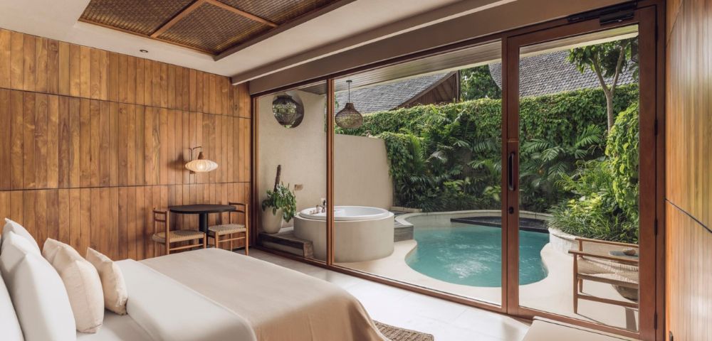 A serene bedroom with a wooden accent wall and soft beige bedding opens to a lush patio featuring a round jacuzzi and a small pool, surrounded by greenery.