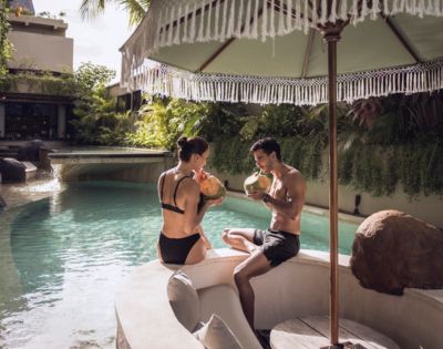 A couple in swimsuits relax by a tropical pool, sipping coconuts under a fringed umbrella. Luxurious, serene setting with lush greenery and a tranquil vibe.