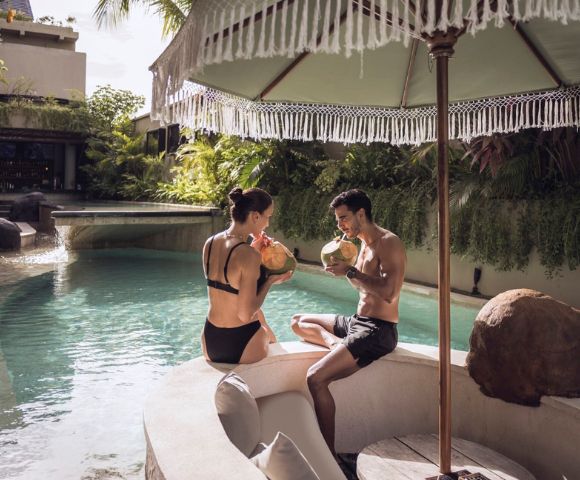 A couple in swimsuits relax by a tropical pool, sipping coconuts under a fringed umbrella. Luxurious, serene setting with lush greenery and a tranquil vibe.