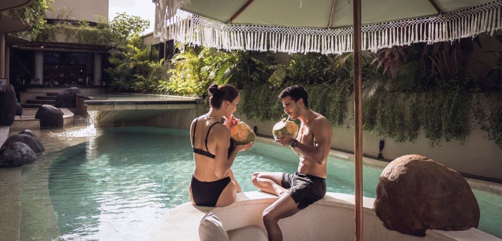 A couple in swimsuits relax by a tropical pool, sipping coconuts under a fringed umbrella. Luxurious, serene setting with lush greenery and a tranquil vibe.