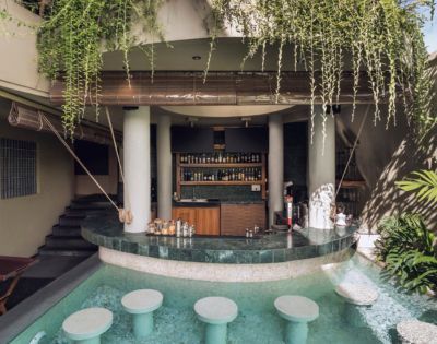Poolside bar with submerged stools in turquoise water. White columns support a roof adorned with lush green vines, creating a tranquil, tropical ambiance.