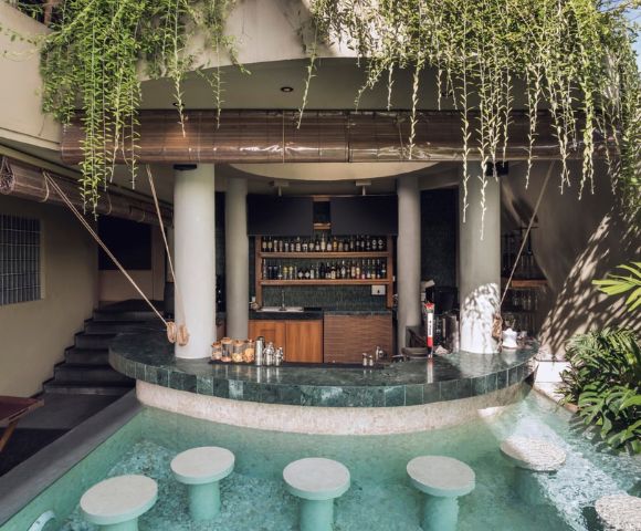 Poolside bar with submerged stools in turquoise water. White columns support a roof adorned with lush green vines, creating a tranquil, tropical ambiance.