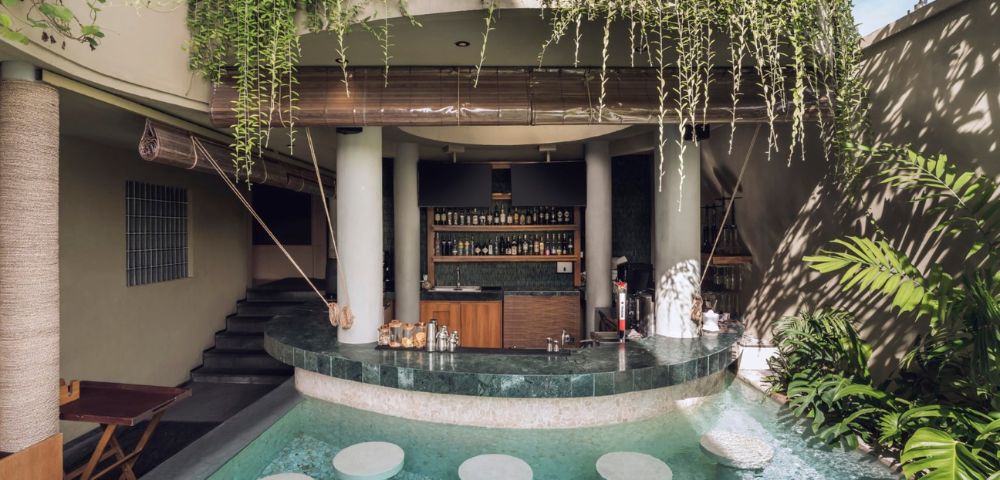 Poolside bar with submerged stools in turquoise water. White columns support a roof adorned with lush green vines, creating a tranquil, tropical ambiance.