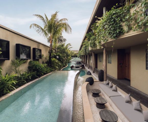 Modern patio with a narrow pool lined by lush plants and palm trees. Cozy seating with cushions and small tables under vine-covered walls, creating a serene, tropical vibe.