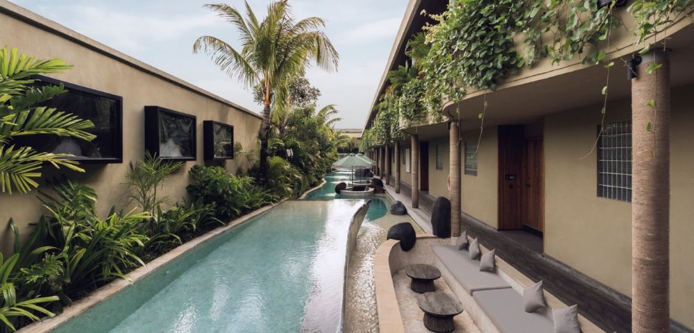 Modern patio with a narrow pool lined by lush plants and palm trees. Cozy seating with cushions and small tables under vine-covered walls, creating a serene, tropical vibe.