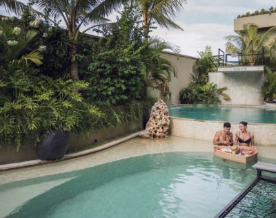 Two people enjoy breakfast on a floating tray in a tranquil pool surrounded by lush tropical plants, creating a serene and luxurious atmosphere.