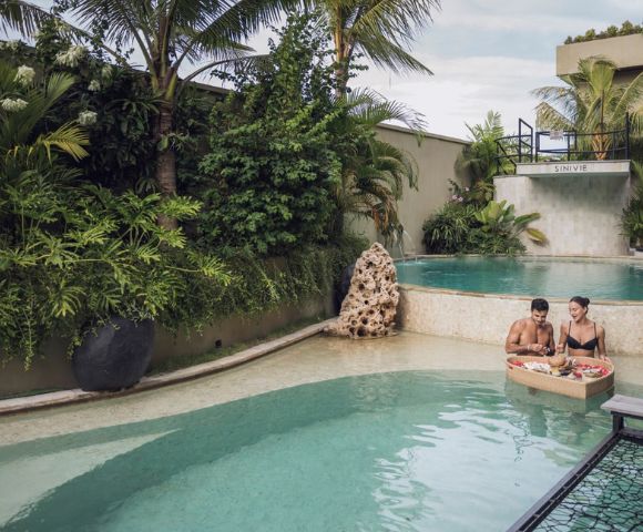 Two people enjoy breakfast on a floating tray in a tranquil pool surrounded by lush tropical plants, creating a serene and luxurious atmosphere.