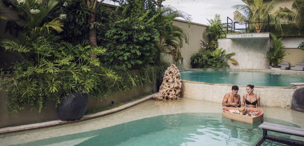Two people enjoy breakfast on a floating tray in a tranquil pool surrounded by lush tropical plants, creating a serene and luxurious atmosphere.