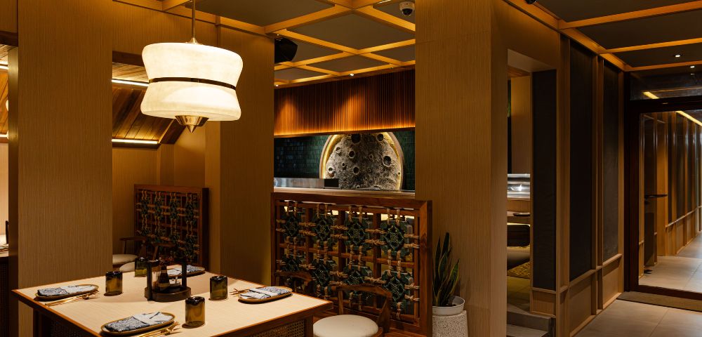 Cozy upscale restaurant interior featuring warm lighting, a wooden table set for dining, elegant pendant lights, and lush greenery accents.