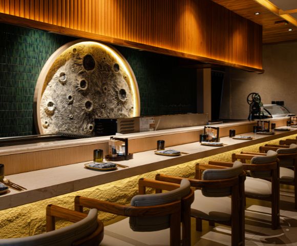 A modern, elegant sushi bar with wooden chairs, a textured circular wall art resembling the moon, warm lighting, and a minimalist design.
