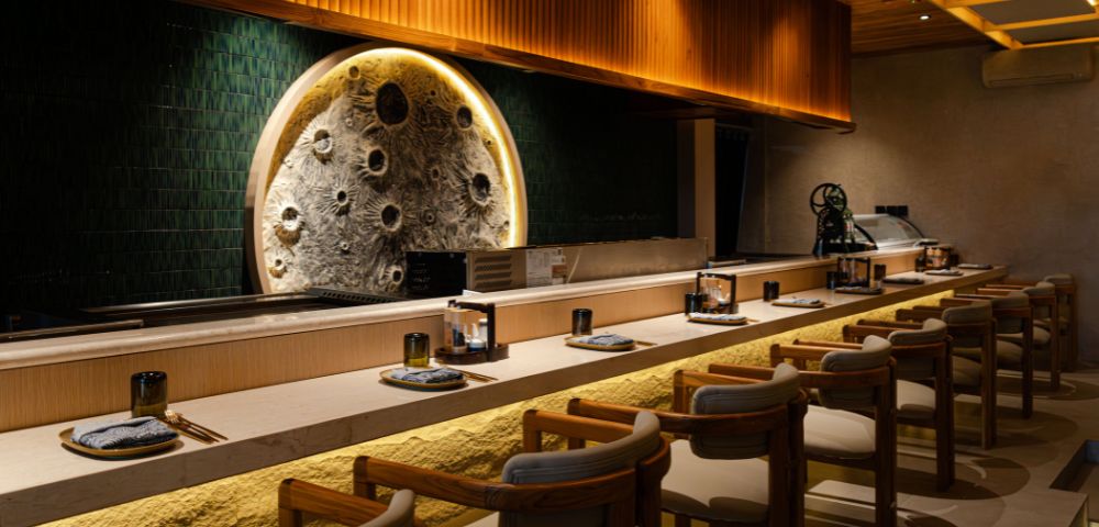 A modern, elegant sushi bar with wooden chairs, a textured circular wall art resembling the moon, warm lighting, and a minimalist design.