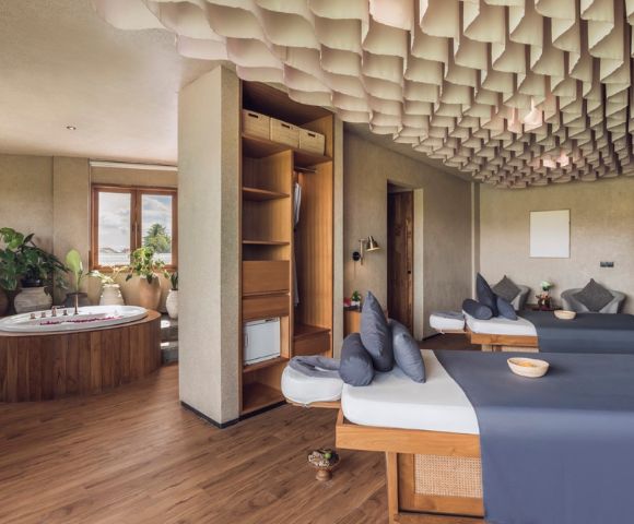 Spacious spa room with two massage tables draped in gray linens, a Jacuzzi by the window, wood flooring, decorative ceiling, and lush plants. Calm ambiance.