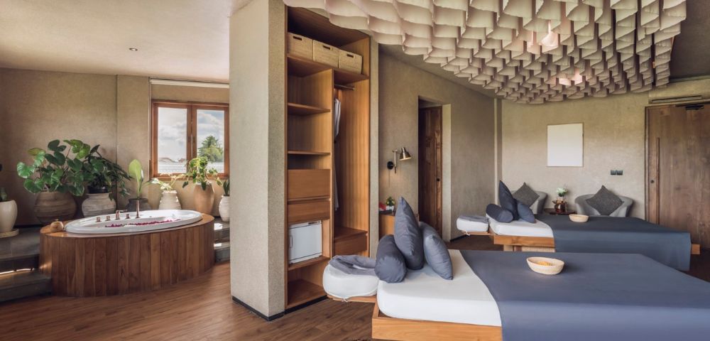 Spacious spa room with two massage tables draped in gray linens, a Jacuzzi by the window, wood flooring, decorative ceiling, and lush plants. Calm ambiance.