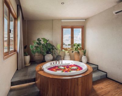 A serene spa room featuring a round wooden hot tub with colorful flower petals, surrounded by lush potted plants, and ambient sunlight from large windows.