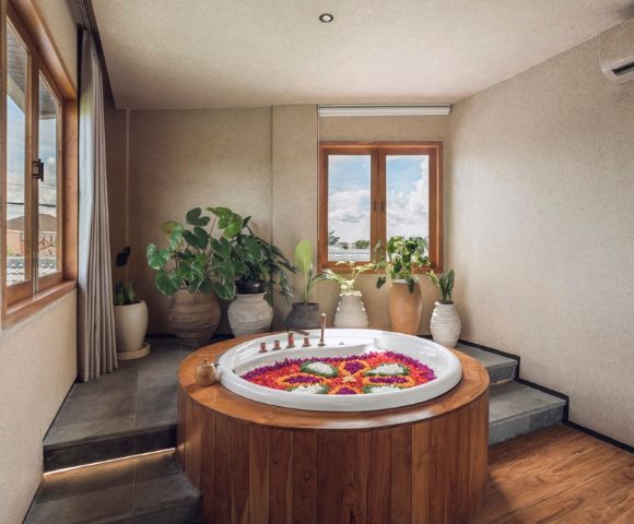 A serene spa room featuring a round wooden hot tub with colorful flower petals, surrounded by lush potted plants, and ambient sunlight from large windows.