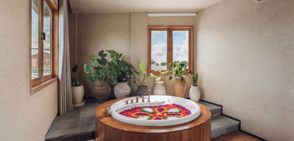 A serene spa room featuring a round wooden hot tub with colorful flower petals, surrounded by lush potted plants, and ambient sunlight from large windows.