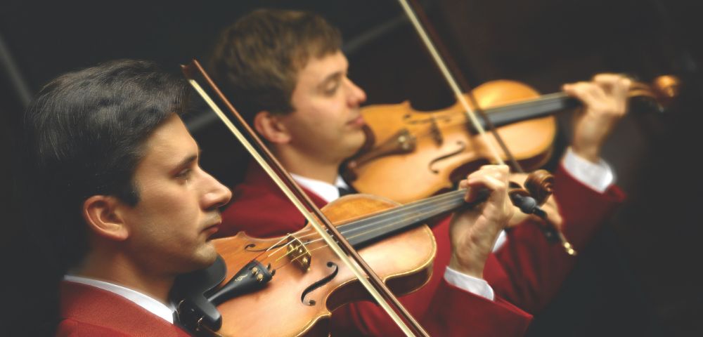 Two musicians in red jackets intensely play violins in an orchestra, creating a focused and harmonious atmosphere. The scene conveys concentration and elegance.