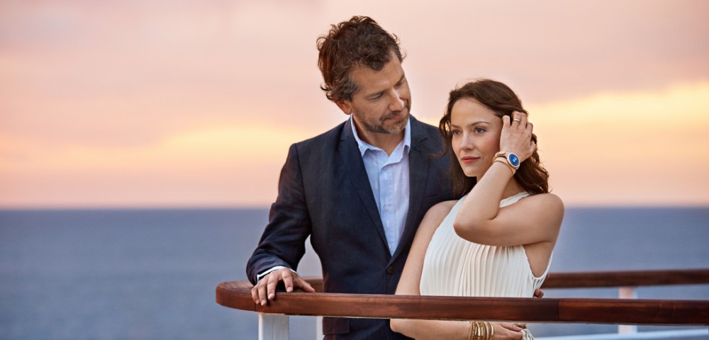 Couple in formal attire standing on a ship deck at sunset, overlooking the ocean with a warm, colorful sky.
